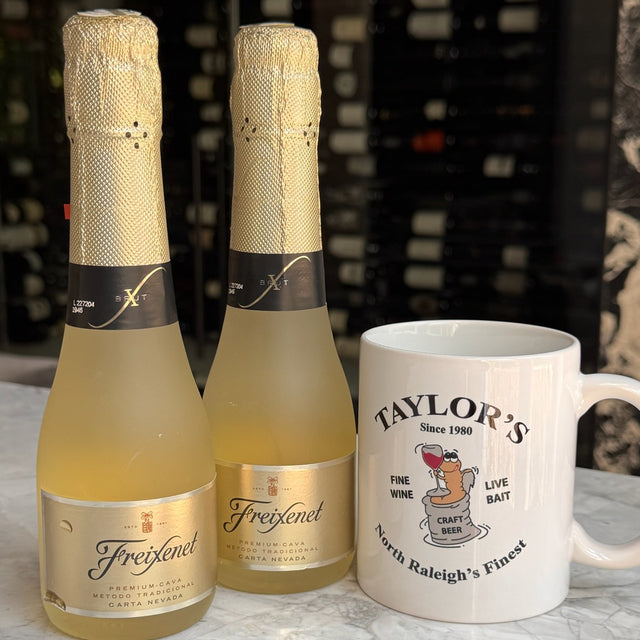 Two bottles of Freyconet sparkling wine and a white ceramic mug printed with 'Taylor's Since 1980' and a cartoon bear, set on a white marble surface with a dark wine cellar background.